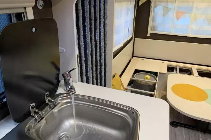 Spüle mit laufendem Wasser in Edelstahlbecken, Armatur, hochgeklappte schwarze Abdeckung neben der Spüle, Vorhang und Fenster dahinter, Sitzbank mit Tisch, gelbe runde Tischunterlage und offene Fächer mit Schüssel