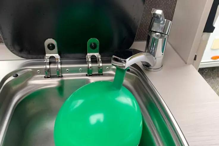 Grüner Ballon in einem Edelstahlspülbecken, Wasser läuft aus einem verchromten Wasserhahn in den Ballon