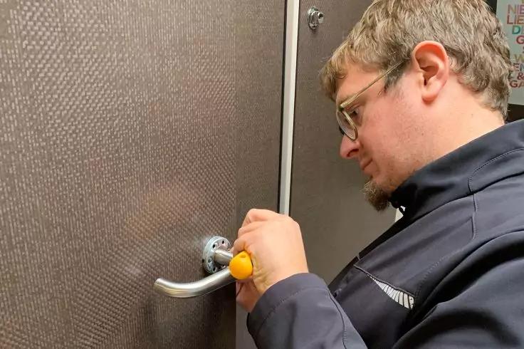 Persoon met bril en donker jack gebruikt een gele schroevendraaier aan een zilveren deurklink van een bruine deur