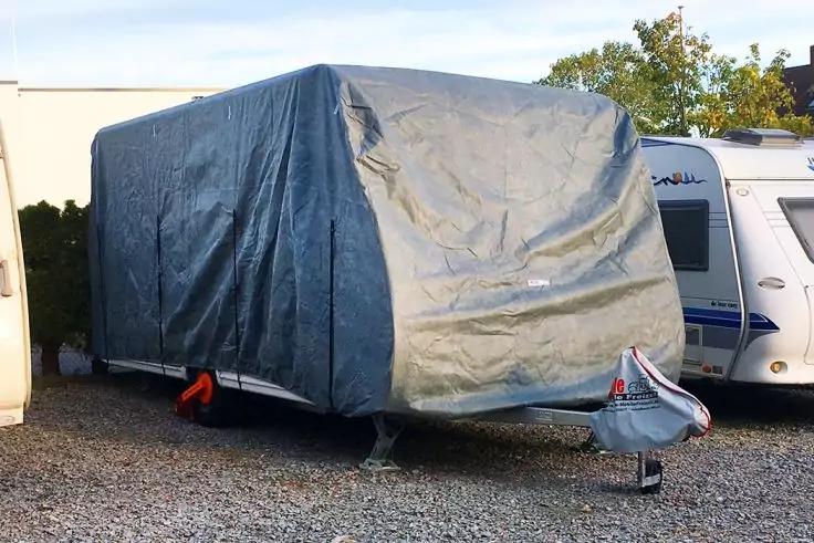 Anhänger mit grauer Plane auf Schotterplatz, Deichsel mit Schutzabdeckung sichtbar, weiße Wohnwagen daneben