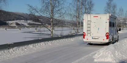 Heckansicht eines wei&szlig;en Wohnmobils auf einer schneebedeckten Stra&szlig;e mit Leitplanke, kahlen Birken, verschneiten H&uuml;geln und blauem Himmel