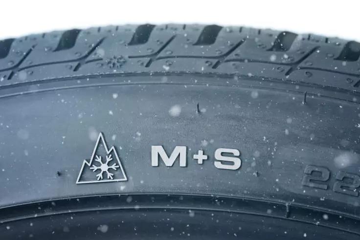 Nahaufnahme der Reifenflanke mit der Aufschrift "M+S" und einem Berg-Symbol mit Schneeflocke, leichte Schneeflocken auf dem Reifen