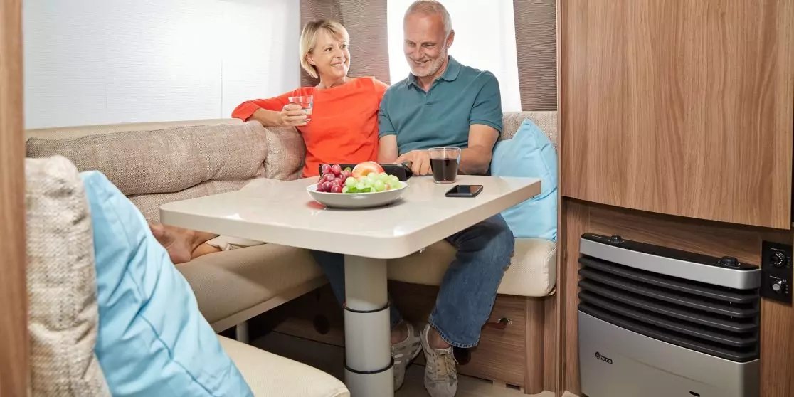 Zwei l&auml;chelnde Personen, Frau in Orange und Mann in Blau, sitzen an einem Tisch im Wohnmobil; Obstschale und Tassen auf dem Tisch, blaue Kissen und Holzschrank sichtbar