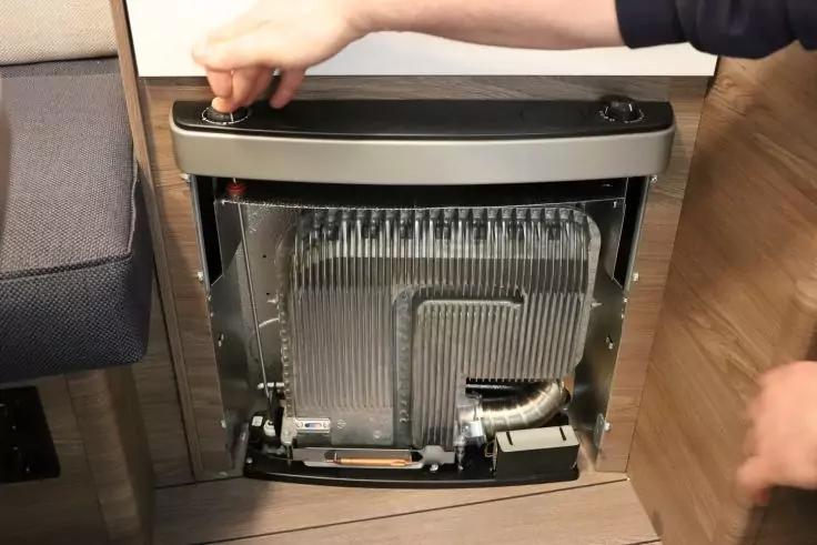 Offenes Metallgehäuse mit lamellenförmigem Metallbauteil, sichtbaren Schläuchen und Kabeln; zwei Hände halten die Klappe; links ein Polstersitz, unten Holzfußboden