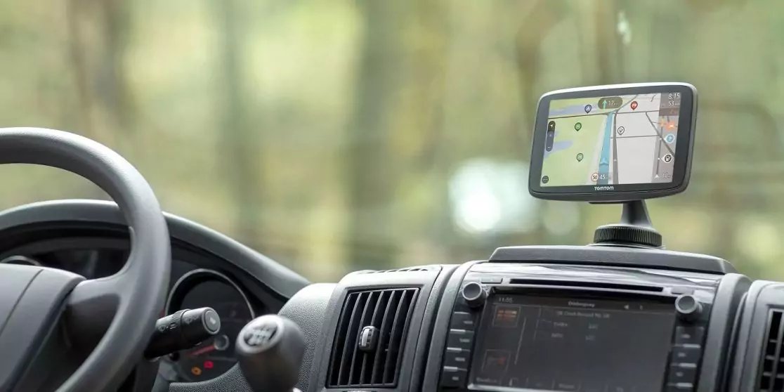 Fahrzeuginnenraum mit links stehenden Lenkrad, Armaturenbrett und mittigem Infotainment; auf dem Armaturenbrett montiertes Navigationsger&auml;t zeigt eine Kartenansicht; durch die Windschutzscheibe sind verschwommene B&auml;ume zu sehen