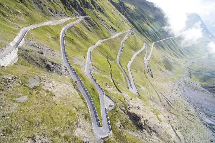 Luftaufnahme einer geschwungenen Bergstra&szlig;e mit mehreren Haarnadelkurven auf einer gr&uuml;nen, felsigen Hangflanke und vereinzelten Wolken