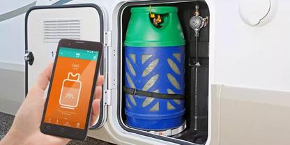 Hand h&auml;lt Smartphone, auf dem eine App 75% F&uuml;llstand anzeigt; daneben in einem Schrankfach eine blau-gr&uuml;ne Gasflasche, mit Gurt gesichert und mit Druckregler