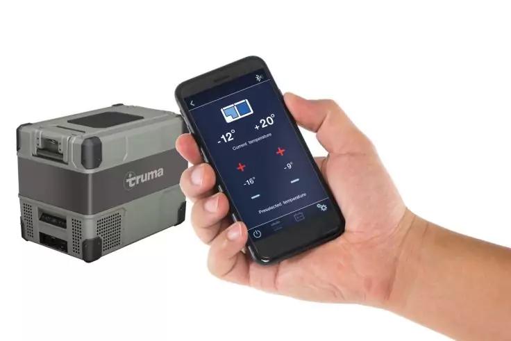 Rechte Hand h&auml;lt Smartphone mit blauem Display, darauf Temperaturzahlen und Symbole; links ein grauer tragbarer K&uuml;hlschrank mit truma-Logo