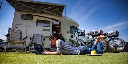 Person liegt auf dem Rasen und benutzt einen Laptop vor einem Wohnmobil; daneben zwei Klappst&uuml;hle, ein Fahrrad und ein Apfel