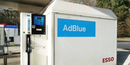 Wei&szlig;es AdBlue-Zapfger&auml;t mit blauem AdBlue-Schild, Zapfpistole und Schlauch, digitalem Display, rotem ESSO-Schriftzug, B&auml;ume und Betonfl&auml;che im Hintergrund
