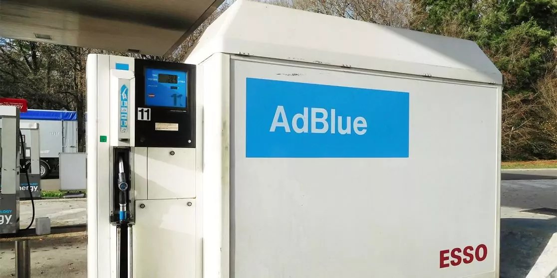 Wei&szlig;es AdBlue-Zapfger&auml;t mit blauem AdBlue-Schild, Zapfpistole und Schlauch, digitalem Display, rotem ESSO-Schriftzug, B&auml;ume und Betonfl&auml;che im Hintergrund