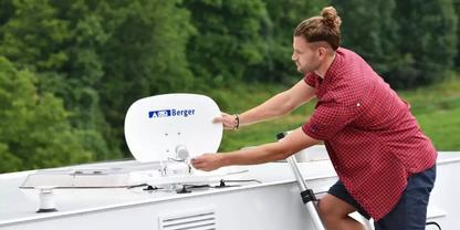 Person in rotem kariertem Hemd kniet auf dem Dach eines wei&szlig;en Fahrzeugs und greift eine wei&szlig;e Satellitensch&uuml;ssel mit 'Berger'-Logo; B&auml;ume im Hintergrund