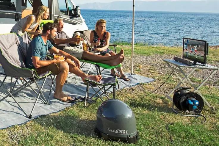 Vier Personen und ein Hund sitzen auf Campingstühlen vor einem Wohnmobil am Meer; ein tragbarer Fernseher auf einem kleinen Tisch, eine Kabeltrommel und eine runde Satellitenkuppel im Vordergrund