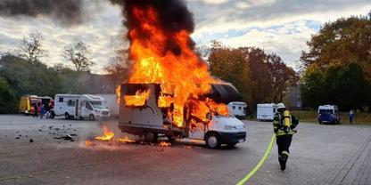 Brennender Wohnwagen mit gro&szlig;en Flammen und schwarzem Rauch auf einem gepflasterten Parkplatz; Feuerwehrperson mit Schlauch n&auml;hert sich; weitere Wohnwagen und B&auml;ume im Hintergrund