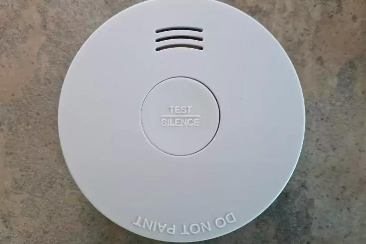 Rundes wei&szlig;es Ger&auml;t mit zentraler Taste 'TEST SILENCE', drei L&uuml;ftungsschlitze oben und Schriftzug 'DO NOT PAINT' an der Kante, grauer Hintergrund