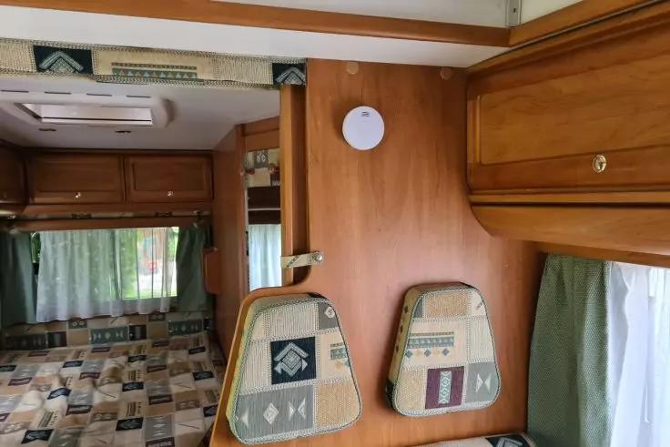 Holzverkleidete Innenwand mit zwei gepolsterten Kissen an einer Trennwand, gemustertes Bett, Fenster mit gr&uuml;nen Vorh&auml;ngen, H&auml;ngeschr&auml;nke, Rauchmelder an der Wand und Deckenl&uuml;ftung