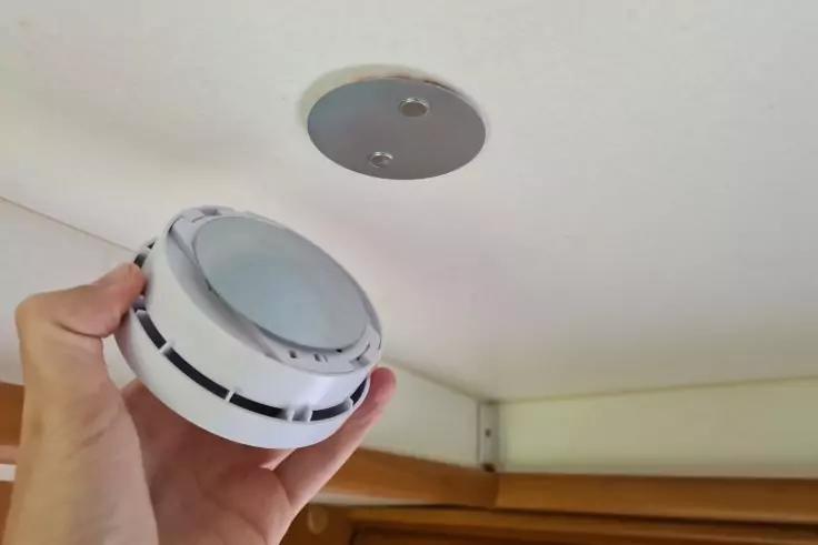 Hand h&auml;lt einen runden wei&szlig;en Rauchmelder nahe einer Decke mit runder Metallhalterung; Kante eines Holzm&ouml;bels sichtbar