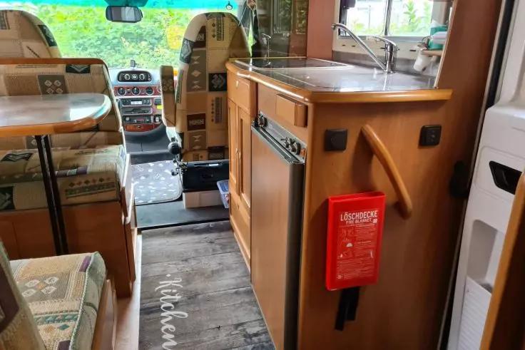 Innenraum eines Wohnmobils: gemusterte Sitzb&auml;nke, kleiner Tisch, Fahrer- und Beifahrersitz, Durchgang zum Fahrerhaus, K&uuml;chenzeile mit Sp&uuml;le und Holzschr&auml;nken, rote Box mit wei&szlig;er Aufschrift an einem Schrank, Bodenmatte mit der Aufschrift 'Kitchen'