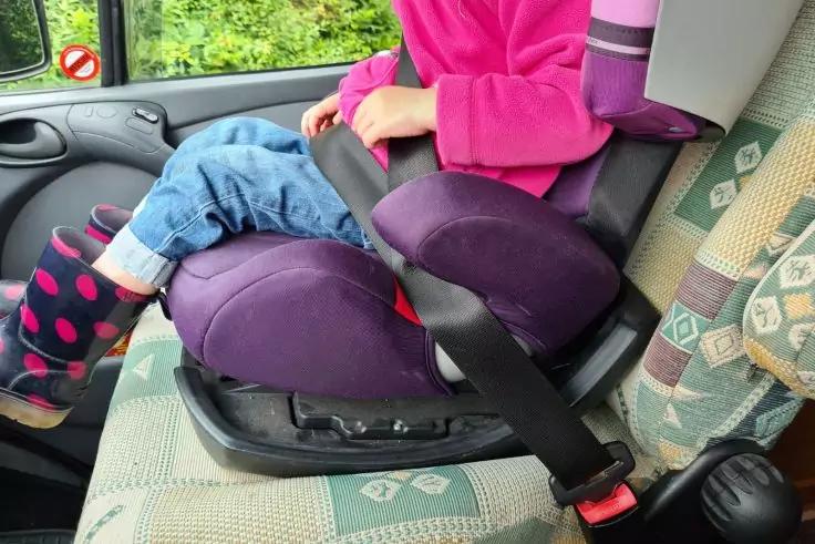 Kind mit pinker Jacke, Jeans und schwarzen Stiefeln mit rosa Punkten sitzt auf einer lila Sitzerh&ouml;hung auf dem Vordersitz eines Autos; Sicherheitsgurt angelegt