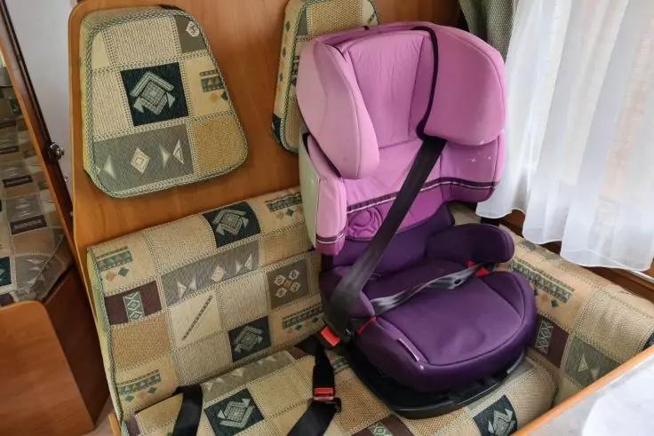 Lila-rosa Kindersitz mit schwarzem Sicherheitsgurt auf gepolstertem, gemustertem Sitz neben einem Fenster mit wei&szlig;em Vorhang