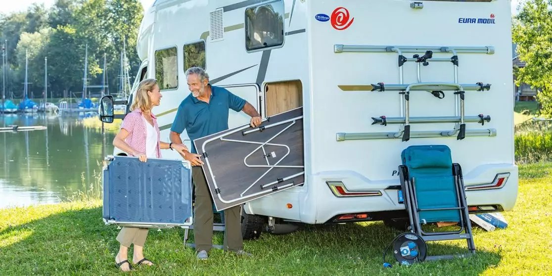 Zwei Personen heben einen klappbaren Tisch aus dem Heckfach eines Wohnmobils; neben dem Wohnmobil steht ein zusammengeklappter Rollstuhl, im Hintergrund ein See mit Booten und B&auml;umen