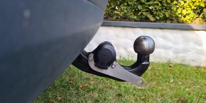 Anh&auml;ngerkupplung mit Kugelkopf und elektrischer Steckdose unter der Hecksto&szlig;stange; Gras, niedrige wei&szlig;e Mauer und Hecke im Hintergrund