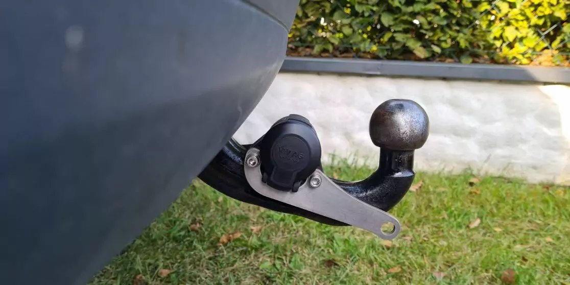 Anh&auml;ngerkupplung mit Kugelkopf und elektrischer Steckdose unter der Hecksto&szlig;stange; Gras, niedrige wei&szlig;e Mauer und Hecke im Hintergrund