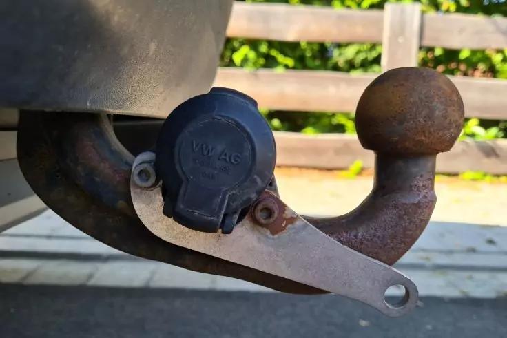 Metallische Anh&auml;ngerkupplung mit Kugel, schwarzer Kunststoffabdeckung an der Steckdose und Rost; Teil des Fahrzeugunterbaus links, Holzzaun im Hintergrund, gepflasterter Boden