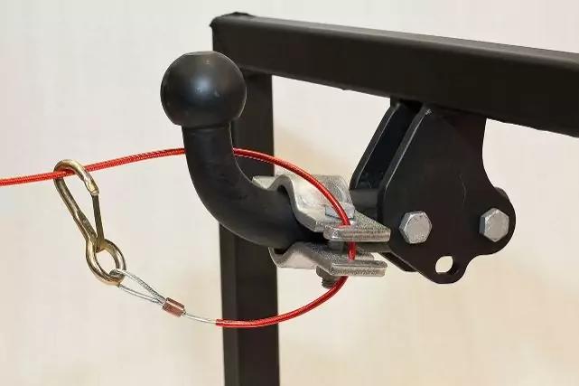 Schwarze Anh&auml;ngerkupplung mit Kugelkopf an einem Metalltr&auml;ger, daran befestigt ein rotes Stahlseil mit Karabiner-Clip und Metallklemme, heller Hintergrund