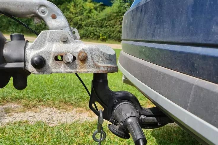 Nahaufnahme einer Metall-Anh&auml;ngerkupplung mit Verriegelung und Rostflecken, angeschlossenem Kabelstecker und sichtbarer blauer Fahrzeugseite; im Hintergrund Gras und B&auml;ume