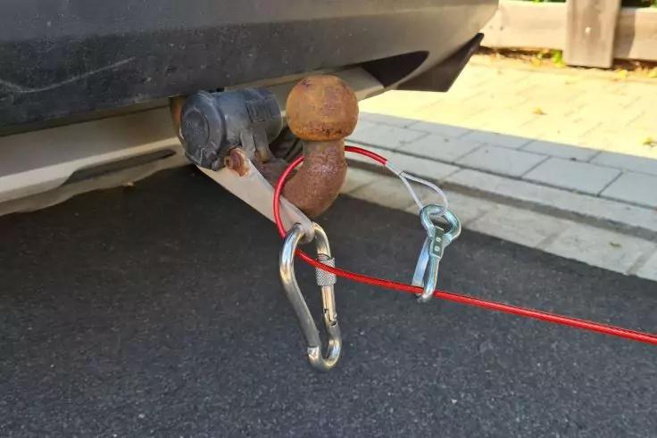 Unterer Bereich eines Autos mit einer rostigen Anh&auml;ngerkupplungskugel, zwei daran befestigten Karabinerhaken und einem roten Kabel; Asphalt und Gehweg im Hintergrund