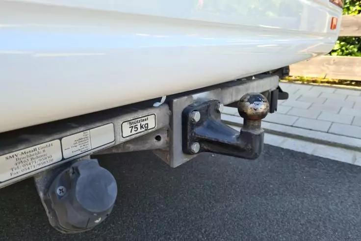 Nahaufnahme der hinteren Anh&auml;ngerkupplung eines wei&szlig;en Autos mit Anh&auml;ngerkugel, elektrischem Steckanschluss, Aufkleber mit 75 kg; Gehweg und Asphalt im Hintergrund