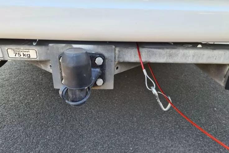 Unterer Bereich der hinteren Sto&szlig;stange eines Fahrzeugs mit Metalltr&auml;ger, schwarzer Steckdosenabdeckung, rotem Sicherungsseil mit Metallkarabiner, kleinem Aufkleber 75 kg und Asphalt darunter