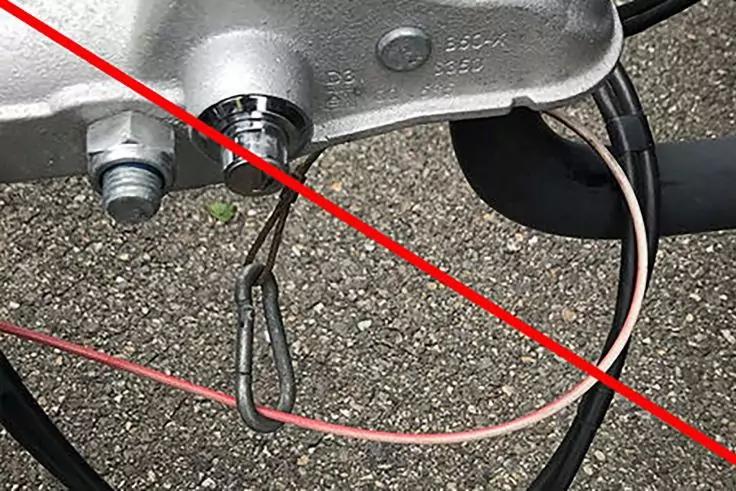 Nahaufnahme einer Metall-Anh&auml;ngerkupplung mit Metallbolzen, Drahtsicherung, rosa und schwarzem Kabel auf kiesigem Untergrund und rotem diagonalen Kreuz