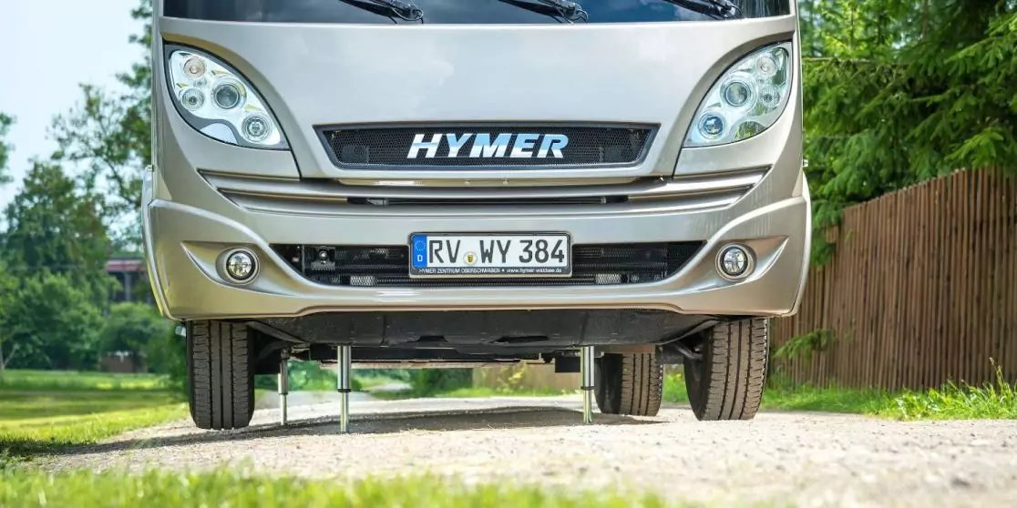 Frontansicht eines Wohnmobils mit Schriftzug 'HYMER' und Kennzeichen 'RV WY 384', auf einem Schotterweg; links B&auml;ume, rechts Holzzaun