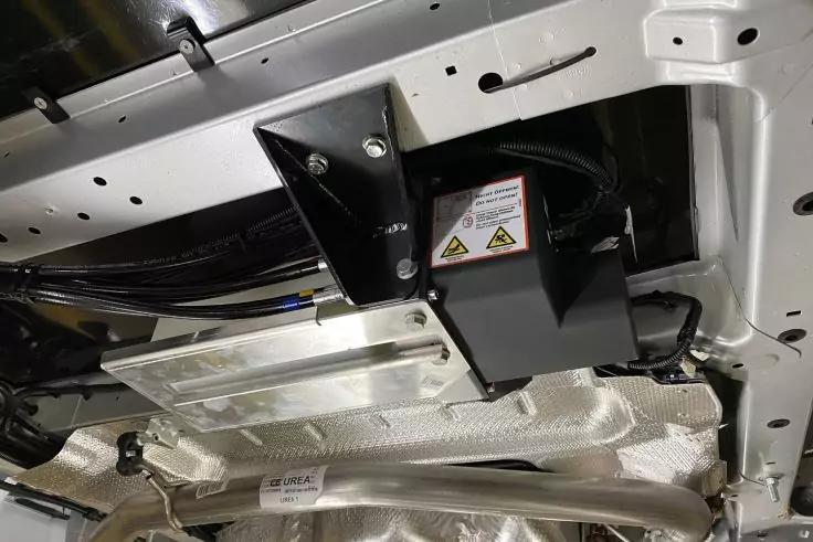 Unterseite eines Fahrzeugs mit wei&szlig;em Metallrahmen, schwarzem rechteckigem Bauteil mit gelben Warnsymbolen, silberner Hitzeschutzfolie, Metallrohren und Kabeln