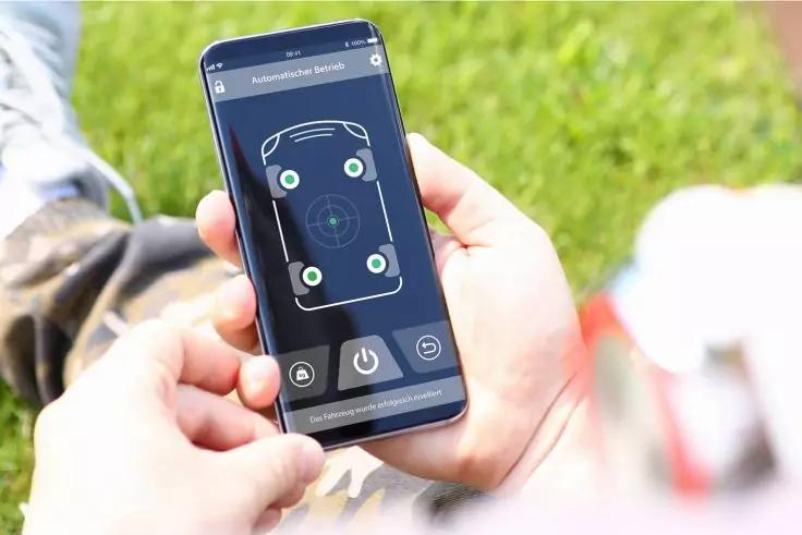H&auml;nde halten ein Smartphone mit einer App-Anzeige: blaues Display mit schematischem Telefon, vier gr&uuml;nen Markierungen und Fadenkreuz in der Mitte; verschwommener Rasen im Hintergrund
