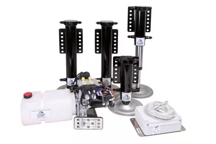 Vier schwarze vertikale S&auml;ulen mit runden Metallf&uuml;&szlig;en, ein wei&szlig;er Kunststoffbeh&auml;lter mit rotem Verschluss, ein Steuerger&auml;t mit Kabeln und Kn&ouml;pfen, eine kleine graue Fernbedienung, eine aufgewickelte wei&szlig;e Kabelrolle und ein flaches graues Bediengeh&auml;u