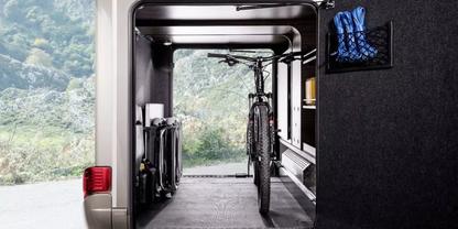 Offenes Heck eines Fahrzeugs mit im Inneren stehendem Fahrrad, Regale an der Seitenwand, blaues Tuch h&auml;ngt an der Wand, gr&uuml;nes H&uuml;gelpanorama durch die vordere &Ouml;ffnung sichtbar