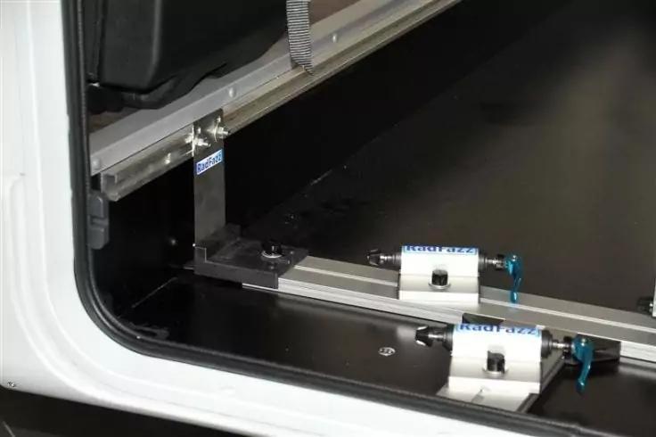 Schwarzer Boden mit einer l&auml;ngs verlaufenden Metallschiene, zwei wei&szlig;en Klemmhaltern mit blauen Griffen auf der Schiene, einem vertikalen Metallwinkel und einem wei&szlig;en Rahmen am linken Bildrand