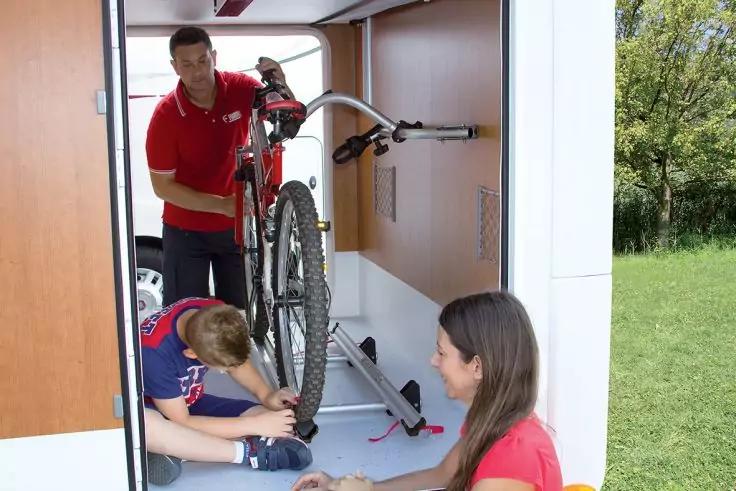 Mann steht in einem Anh&auml;nger und h&auml;lt ein Fahrrad; Junge kniet und arbeitet am Vorderrad; Frau sitzt auf der Schwelle; Gras rechts sichtbar