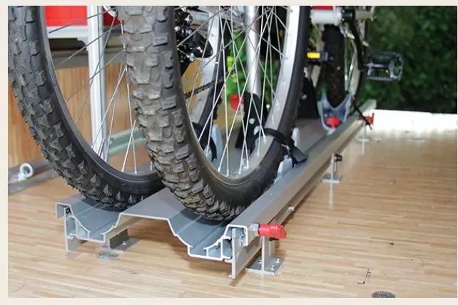 Zwei Fahrradreifen mit grobem Profil in einer Metallschiene auf Holzboden; Teile von Fahrradrahmen und rote Klemmen sichtbar