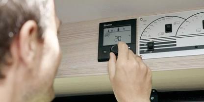 Hand dreht an einem wandmontierten Bedienfeld mit digitaler Anzeige '20', daneben analoge Drehskalen; unscharfer Kopf einer Person links, helles Holzpaneel im Hintergrund