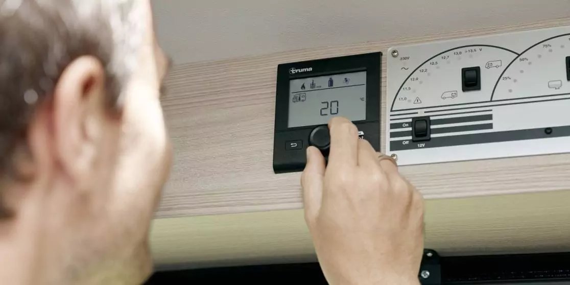 Hand dreht an einem wandmontierten Bedienfeld mit digitaler Anzeige '20', daneben analoge Drehskalen; unscharfer Kopf einer Person links, helles Holzpaneel im Hintergrund