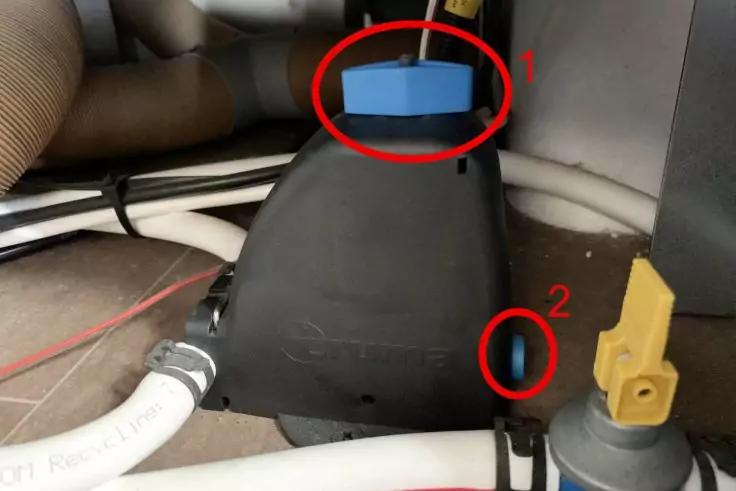 Innenraum eines Schranks mit schwarzem Kunststoffgeh&auml;use, wei&szlig;em Schlauch links, blauem rechteckigem Bauteil oben, rotem Kreis mit Ziffer 1, kleinem seitlichem Rundloch mit rotem Kreis und Ziffer 2, gelbem Kunststoffventil rechts und Kabelbindern