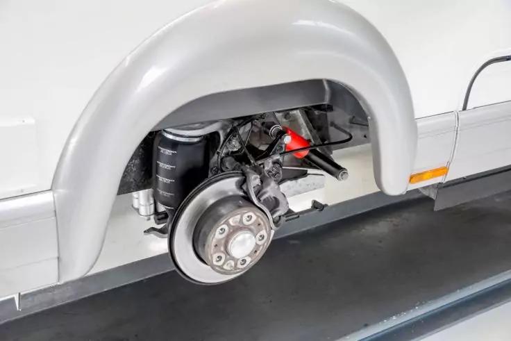 Freigelegter Radbereich eines wei&szlig;en Fahrzeugs ohne Reifen, sichtbare Bremsscheibe, roter Bremssattel und Federbein im Radkasten