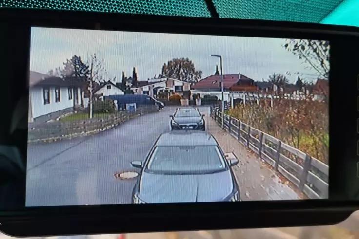 Blick durch ein rechteckiges Display auf eine Wohnstraße mit zwei geparkten Autos, Gehweg, Holzgeländer rechts, Einfamilienhäuser, Bäume und eine Straßenlaterne