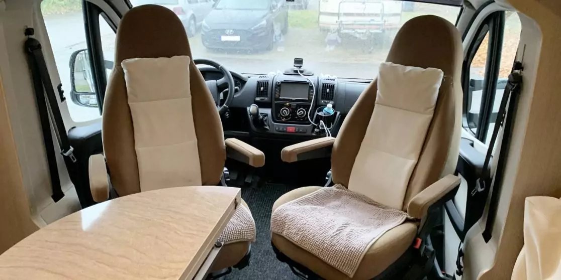 Zwei beige Sitze vorne, Lenkrad und Armaturenbrett, links ein Tisch, durch die Windschutzscheibe geparkte Autos sichtbar
