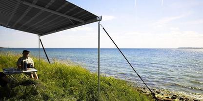 Person sitzt auf einem Campingstuhl unter einem rechteckigen dunklen Dach, daneben ein kleiner Campingtisch mit Kanne und Tasse, gr&uuml;ne Uferb&ouml;schung mit Gras und Steinen, im Hintergrund Meer und Himmel