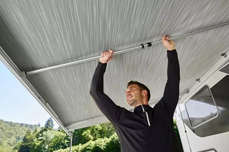 Mann in dunklem Langarmshirt hält mit beiden Händen die Unterseite einer ausziehbaren Wohnmobilmarkise; rechts das Wohnmobil, im Hintergrund Bäume und Hügel